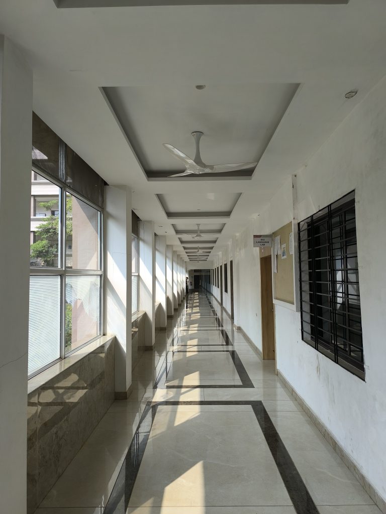 A long, sunlit corridor with large windows on the left, allowing natural light to create patterns on the polished floor. The ceiling features modern fans, and there are bulletin boards and doors on the right wall. The corridor extends into the distance.