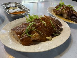 Juicy roasted duck pieces served on white plates with green onion garnish and dipping sauces in a metal container, showing the glossy brown skin and tender meat underneath.