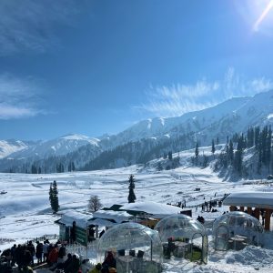 A scenic winter landscape featuring snow-covered mountains and pine trees under a clear blue sky. In the foreground, there are transparent domed structures on a snowy field, with a group of people gathered around, possibly enjoying a snowy outing.