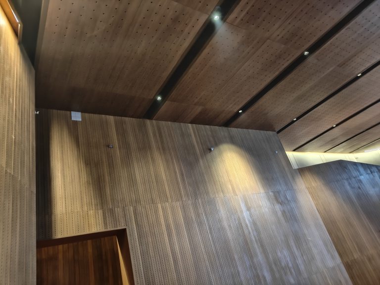 Interior view of a room with wooden paneled walls and ceiling, featuring recessed lighting. The ceiling and walls have a perforated pattern.