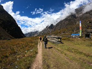 A scenic mountain landscape with a dirt path winding through a grassy valley. Two hikers are walking along the path, surrounded by tall mountains on either side. The sky is vibrant blue with scattered clouds. A long line of stacked stones and prayer flags is visible to the right of the path.