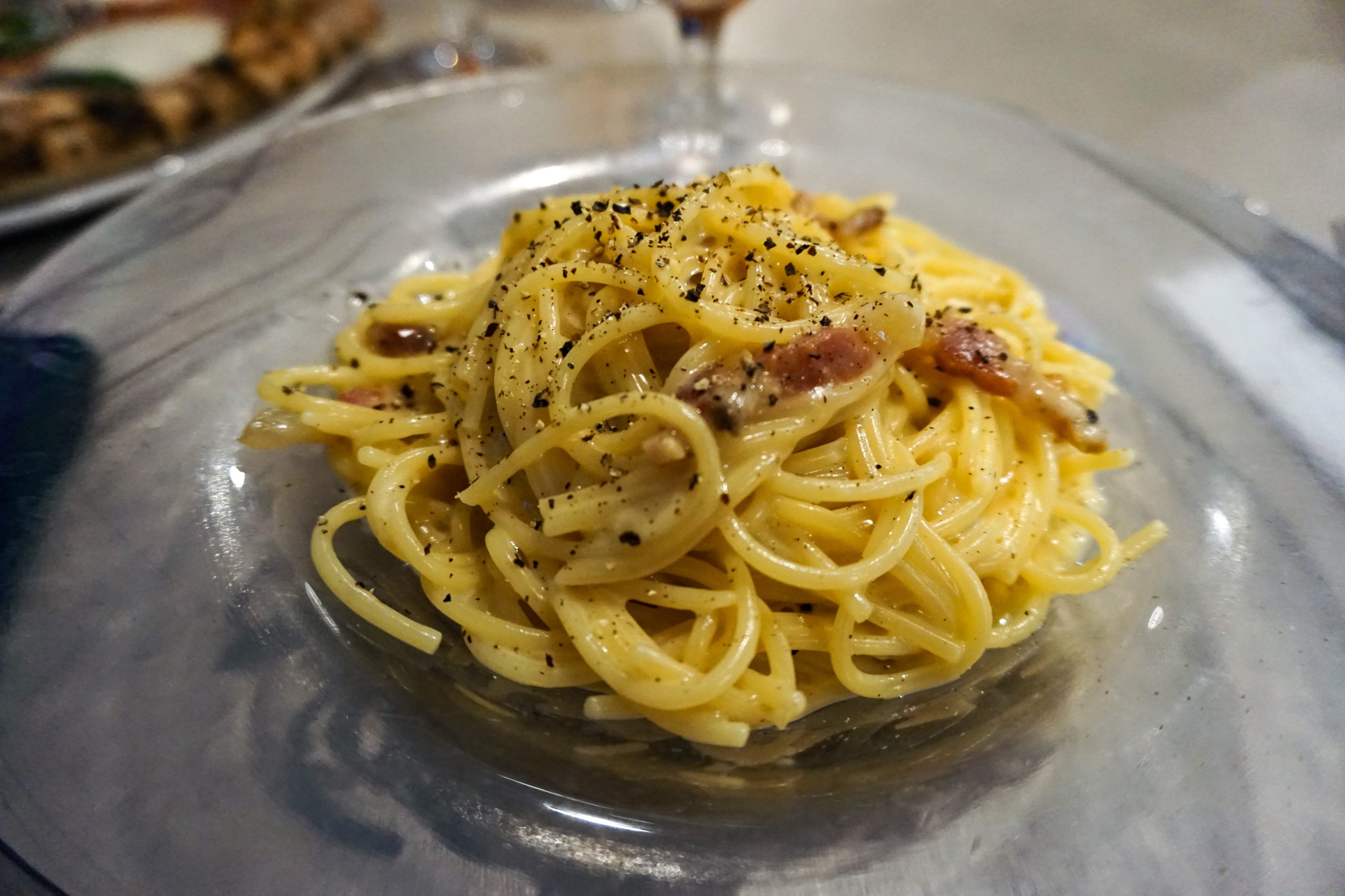 A close-up of a dish of spaghetti alla carbonara, garnished with black pepper and small pieces of guanciale, served on a clear glass plate.