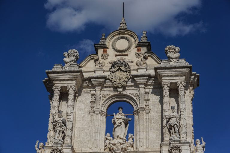 Intricate architectural facade featuring statues and ornate carvings set against a blue sky with clouds.