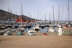 A marina filled with numerous sailboats and yachts docked in calm water. In the background, a large red ship is visible near a mountainous landscape. In the foreground, there are parked cars and part of a wooden promenade.