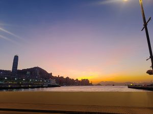 A waterfront cityscape at sunset, featuring a skyline with tall buildings silhouetted against a colorful sky