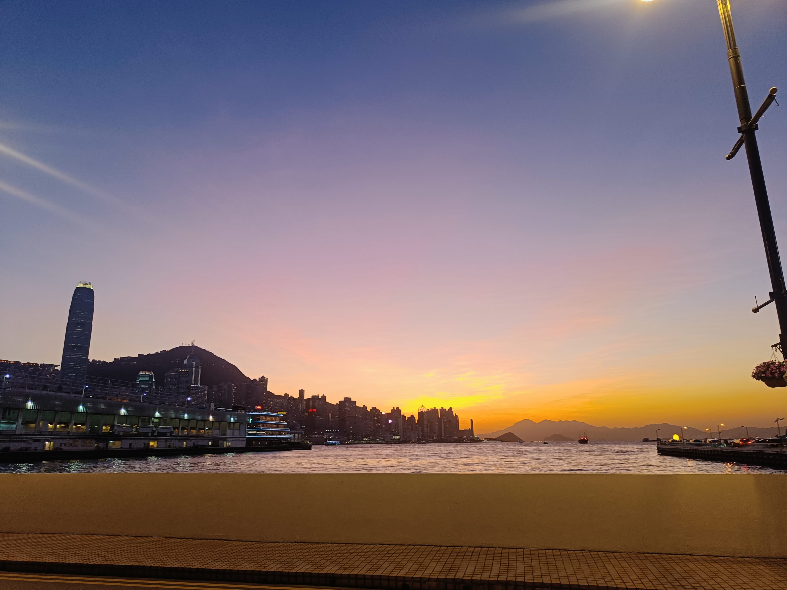 A waterfront cityscape at sunset, featuring a skyline with tall buildings silhouetted against a colorful sky