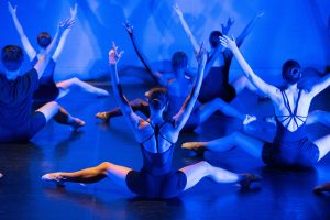 A group of dancers performing on stage under blue lighting, with their arms raised and legs extended, showcasing synchronized movement and grace.