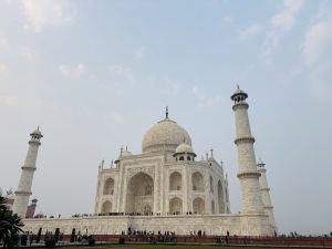 The image shows the Taj Mahal, a large white marble mausoleum with a prominent dome and minarets on each corner, set against a clear sky. There are people gathered along the base of the structure.