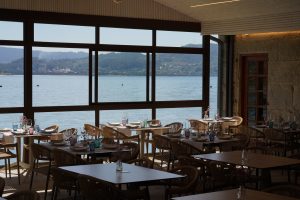 A restaurant with wooden tables and chairs set with colorful glassware and vases with flowers, overlooking a scenic view of a Ria de Pontevedra surrounded by forested hills through large windows.