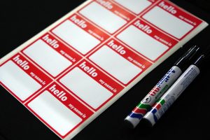A sheet of red and white name tags labeled &quot;hello my name is&quot; next to two black markers, on a black surface.