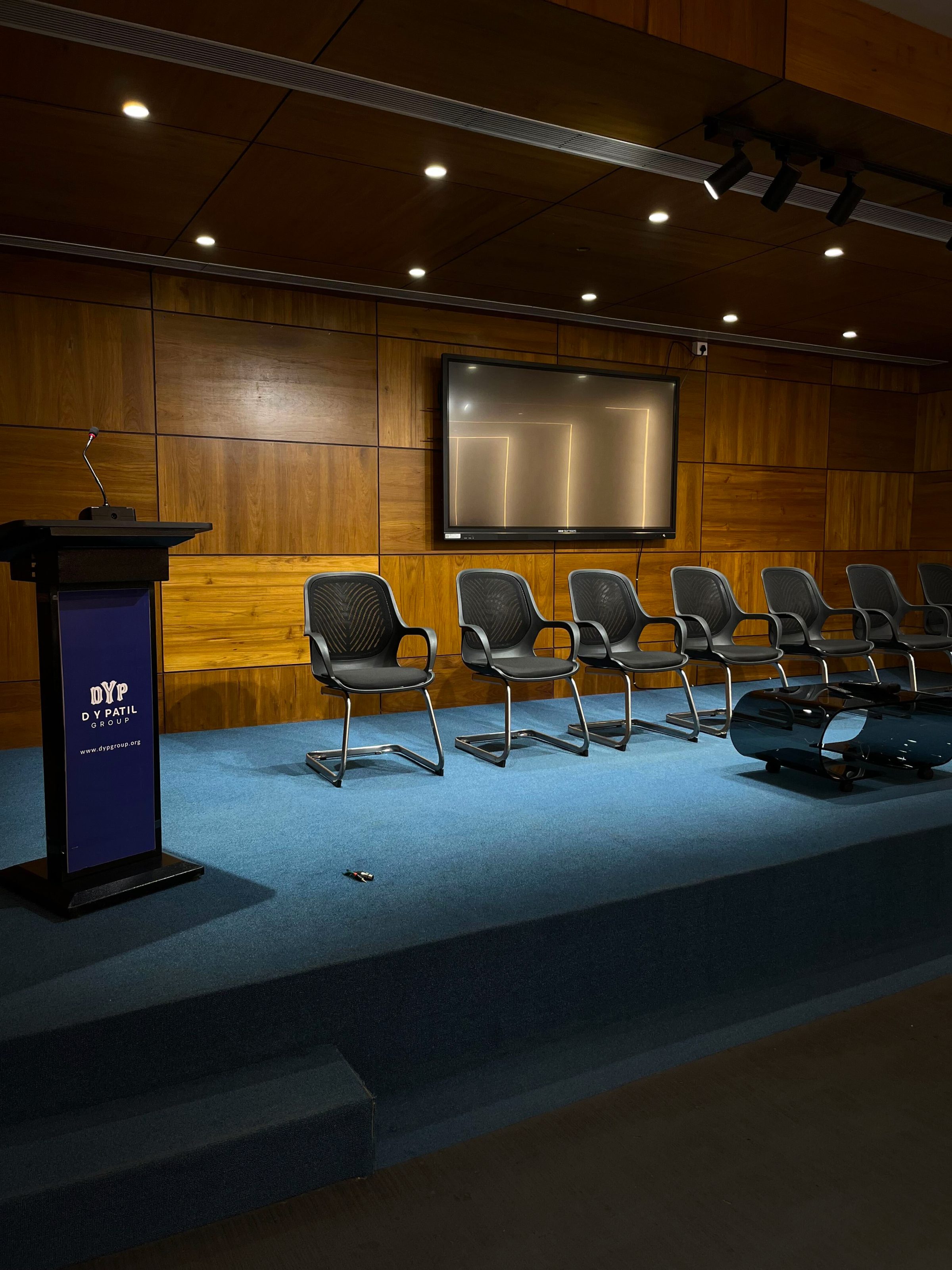 This image depicts a well-organized stage setup in an auditorium or conference hall. The stage features a wooden backdrop with a mounted flat-screen display in the center. 