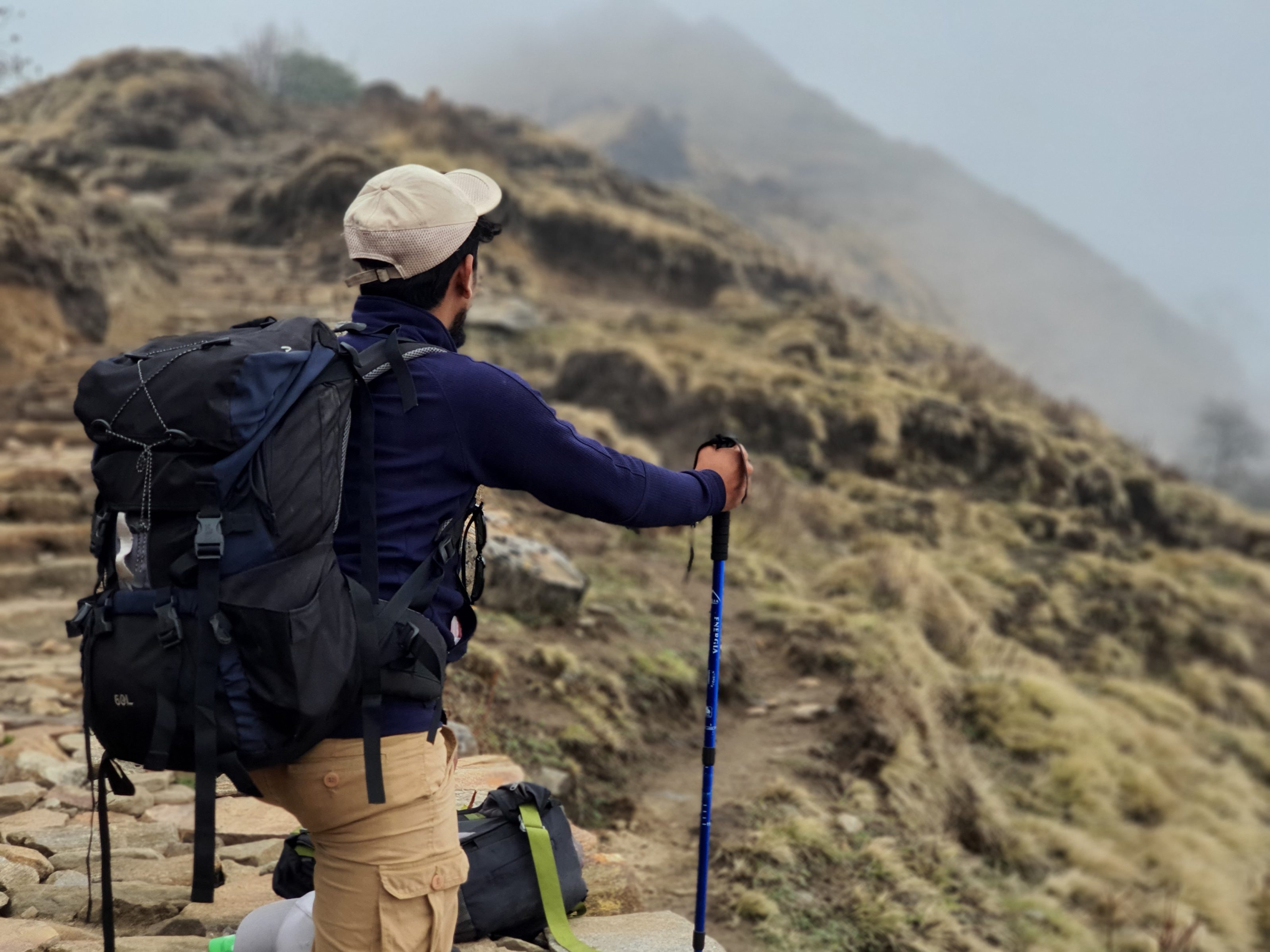 A person wearing a beige cap and carrying a large backpack is standing on a stone path, holding a trekking pole. They are looking at a misty, hilly landscape covered in grass and rocks.