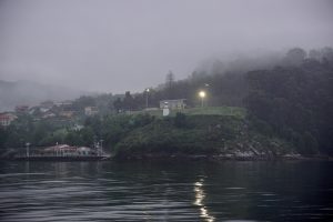 A misty landscape with a grassy hill topped by a building and illuminated streetlamps. The hill is surrounded by lush greenery and overlooks calm water. Houses are visible in the distance against a backdrop of mist-covered trees.