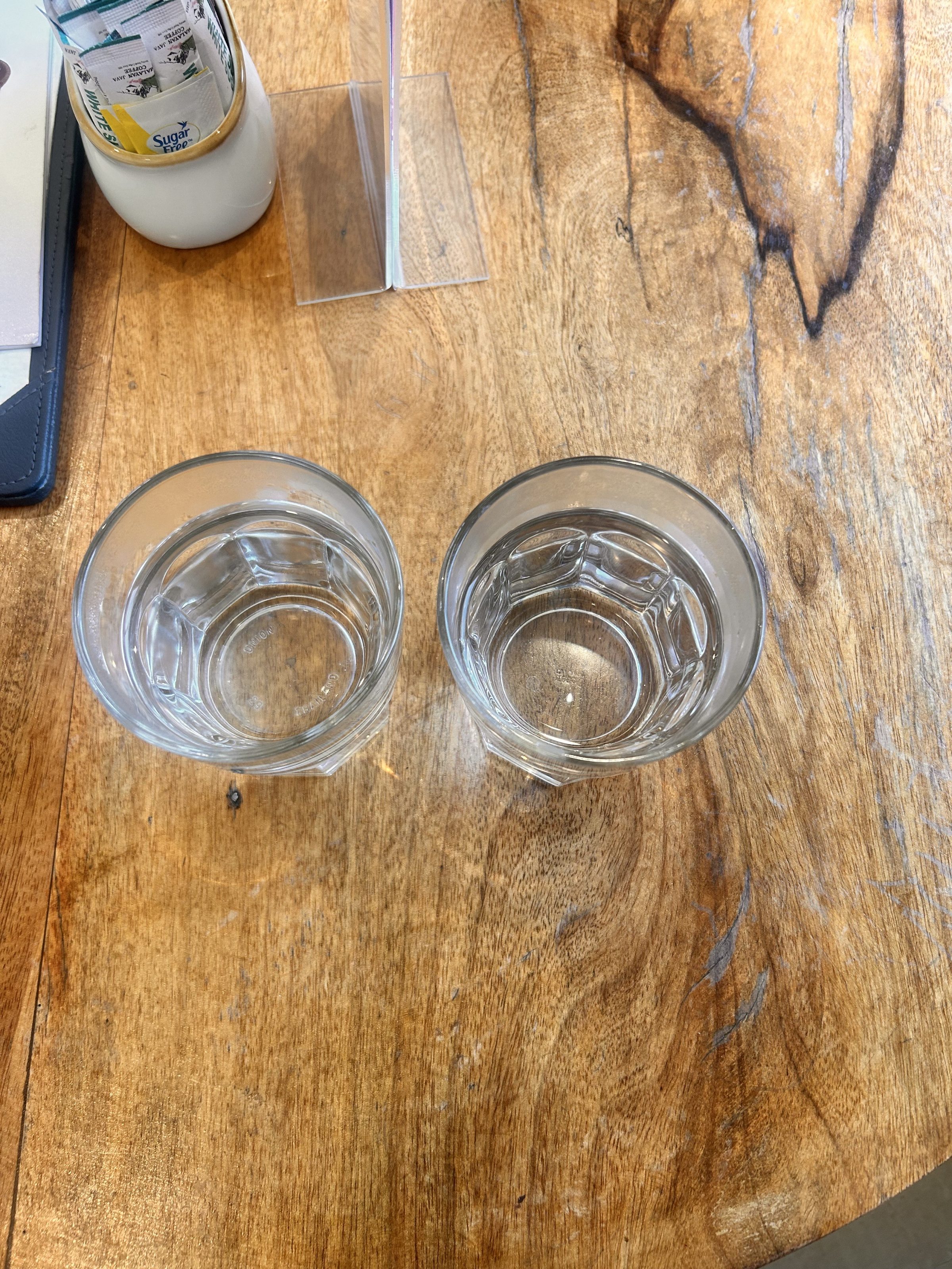 Two glasses of water on a wooden table, with a container holding sugar packets and a clear acrylic menu holder nearby.