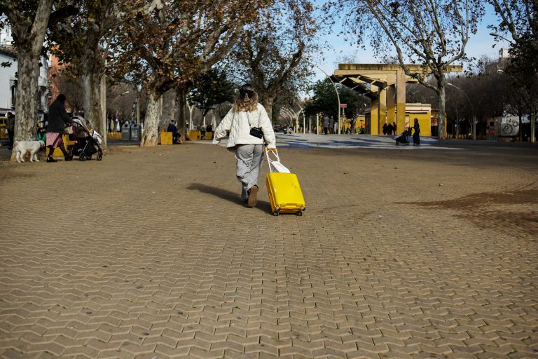 A person walking on a tree-lined paved pathway, pulling a bright yellow suitcase.