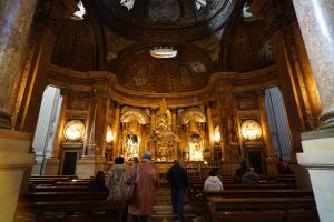 Interior of an ornate church with a richly decorated ceiling and altar, featuring sculptures and paintings. Several people are seated on wooden pews, some standing and observing the artwork.