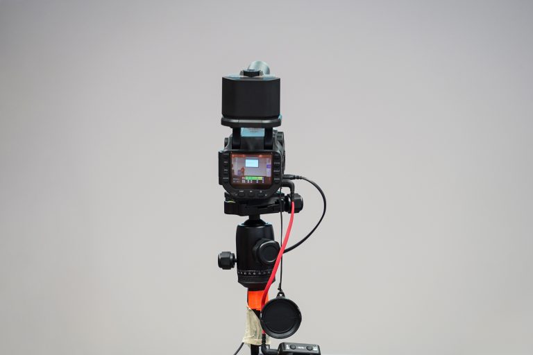 A professional camera setup on a tripod, facing towards the viewer. The camera screen is visible, showing a preview of what the camera is capturing. A black lens cap hangs from the tripod, and several cables are connected to the camera.