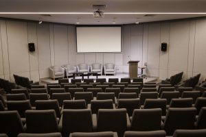 An empty conference room with rows of chairs facing a stage area. There are five gray chairs and two small tables on the stage, with a podium and microphone to the right. A large blank screen is mounted on the wall behind the stage. The room has gray walls and a ceiling-mounted projector.