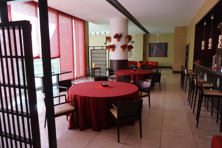 A modern tiled floor dining area with round tables covered in red tablecloths, dark chairs, and a long bar with high stools. Floral arrangements hang on a central white column, and red blind partially cover large windows, allowing natural light to illuminate the space.