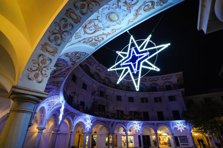 A nighttime view of an ornate, open plaza with decorative arches and illuminated star-shaped christmas lights hanging from the ceiling. The arches are intricately painted with floral and geometric patterns, and a few storefronts are visible below the arches.