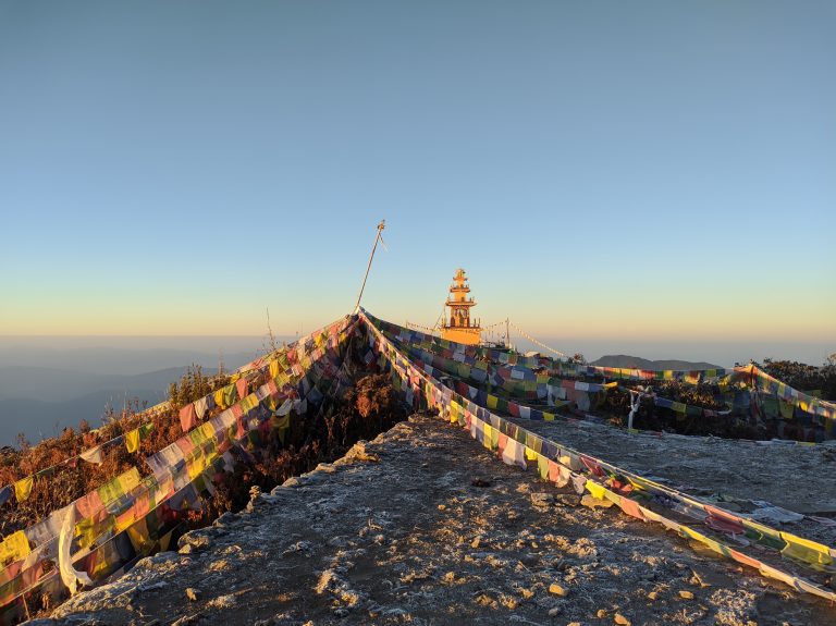 A mountainous landscape at sunrise with colorful prayer flags strung across the foreground. In the distance, there is a multi-tiered structure resembling a stupa, and the sky is clear with a soft gradient from blue to yellow near the horizon.