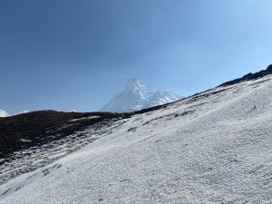 Snow-covered mountain slope with a distant peak visible against a clear blue sky. Sparse vegetation peeks through the snow on the slope.