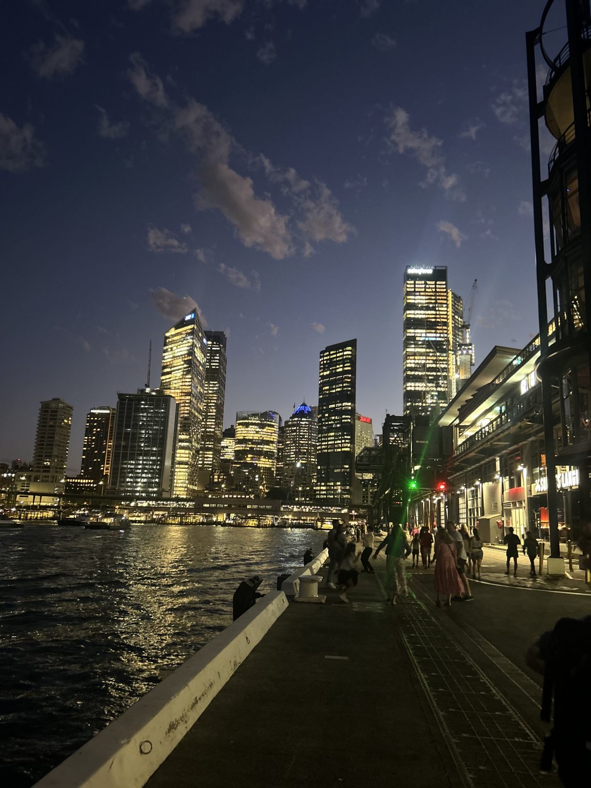 A nighttime cityscape showing illuminated skyscrapers reflecting on a body of water. A walkway runs alongside the water with people walking, and the sky is dotted with clouds as evening sets in.