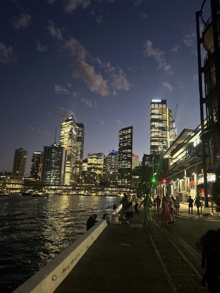 A nighttime cityscape showing illuminated skyscrapers reflecting on a body of water. A walkway runs alongside the water with people walking, and the sky is dotted with clouds as evening sets in.