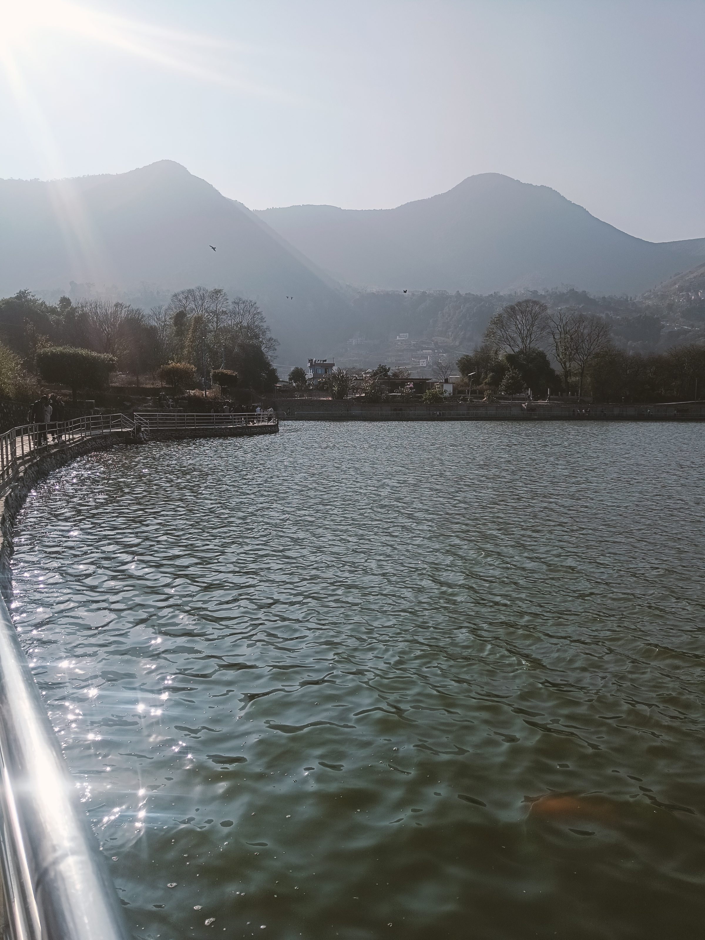 A scenic view of of Taudaha lake near Chobhar region, with sunlight reflecting off its surface, bordered by a railing on the left. In the background, there are distant mountains and a cloudy sky with sunlight streaming through.