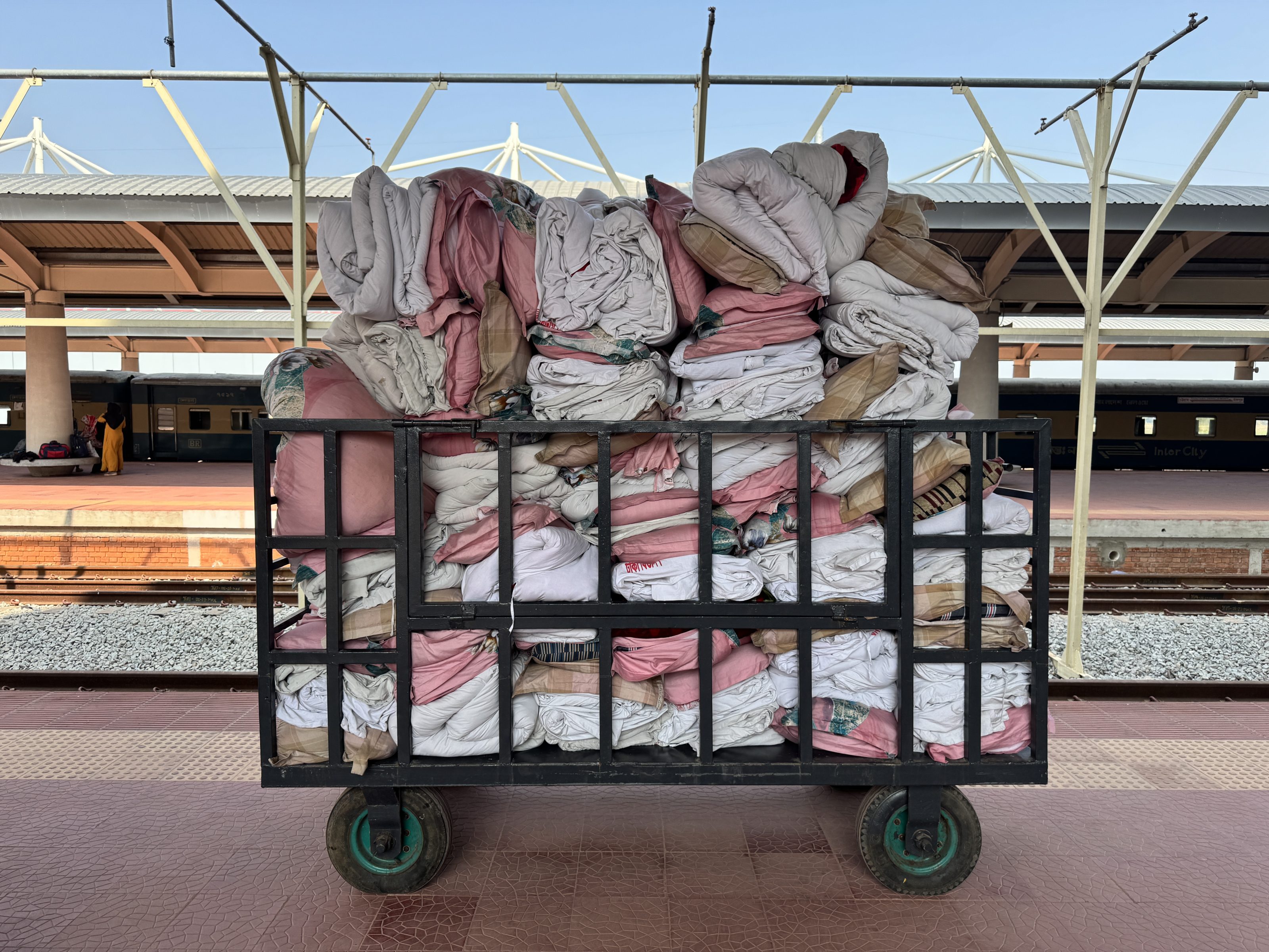 A metal cart loaded with stacks of folded pink and white bedding or linens sits on a train platform, with green wheels and a grid-like frame to hold everything in place.