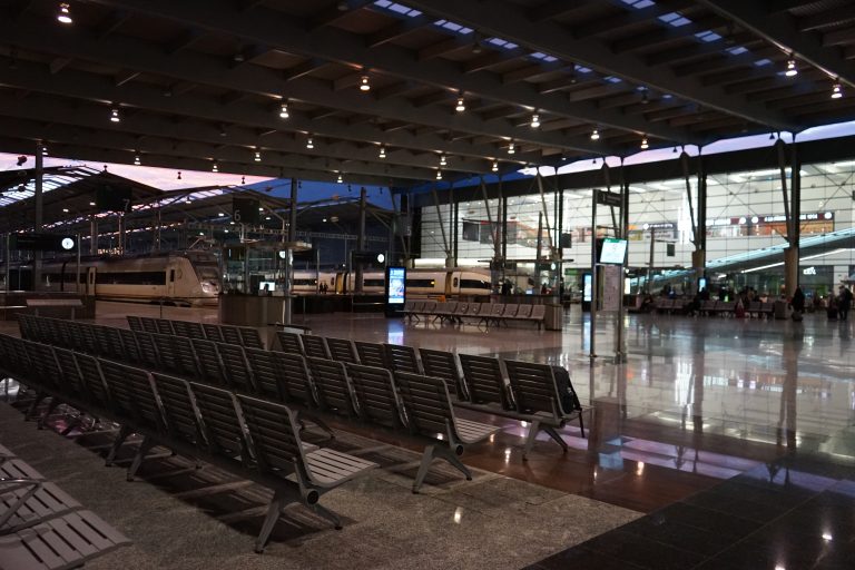 A spacious, modern train station interior with rows of empty seats in the foreground. Several trains are parked at platforms under a large, well-lit roof.