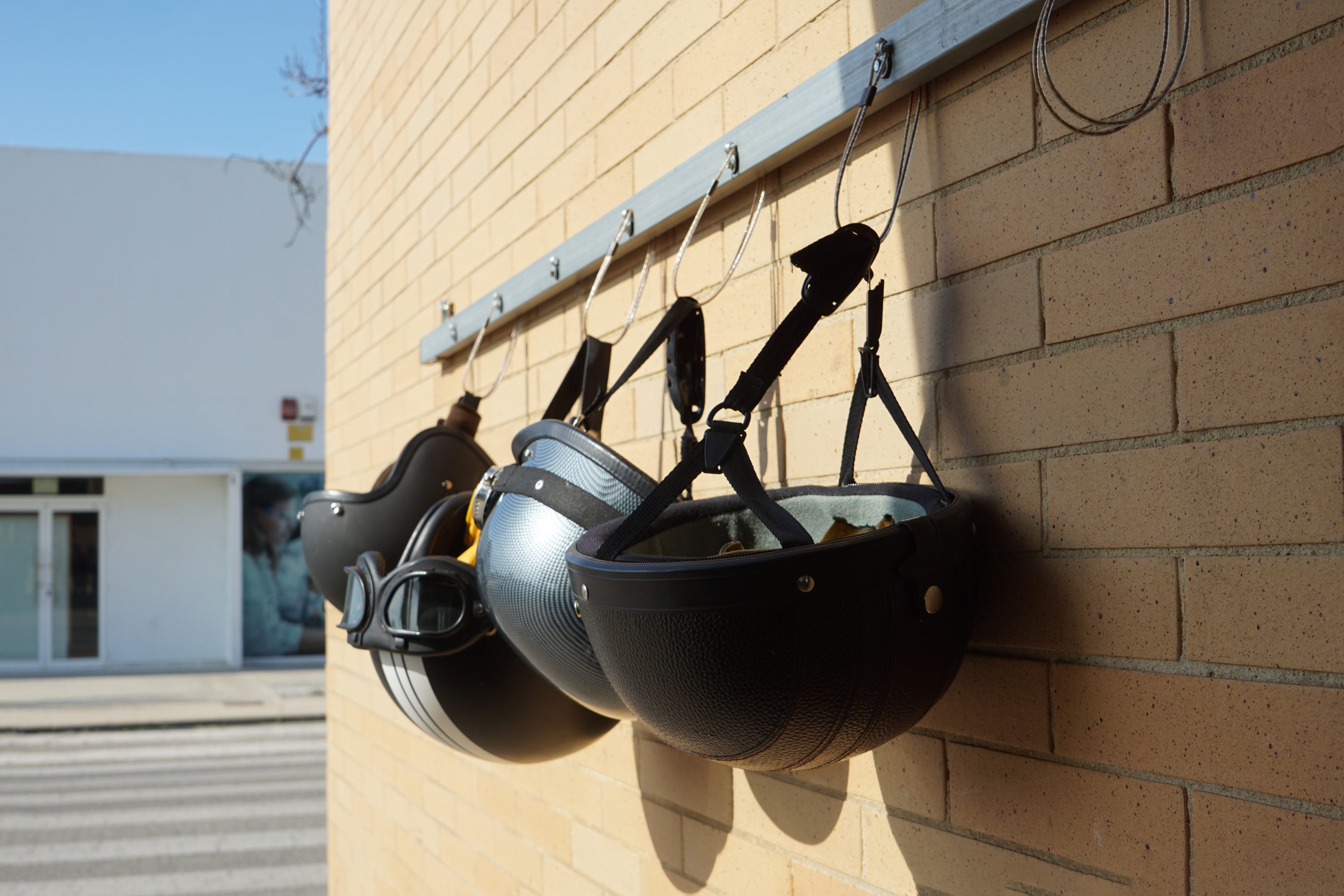 Four motorcycle helmets are hanging on metal hooks attached to a brick wall, with a clear blue sky and a building in the background.