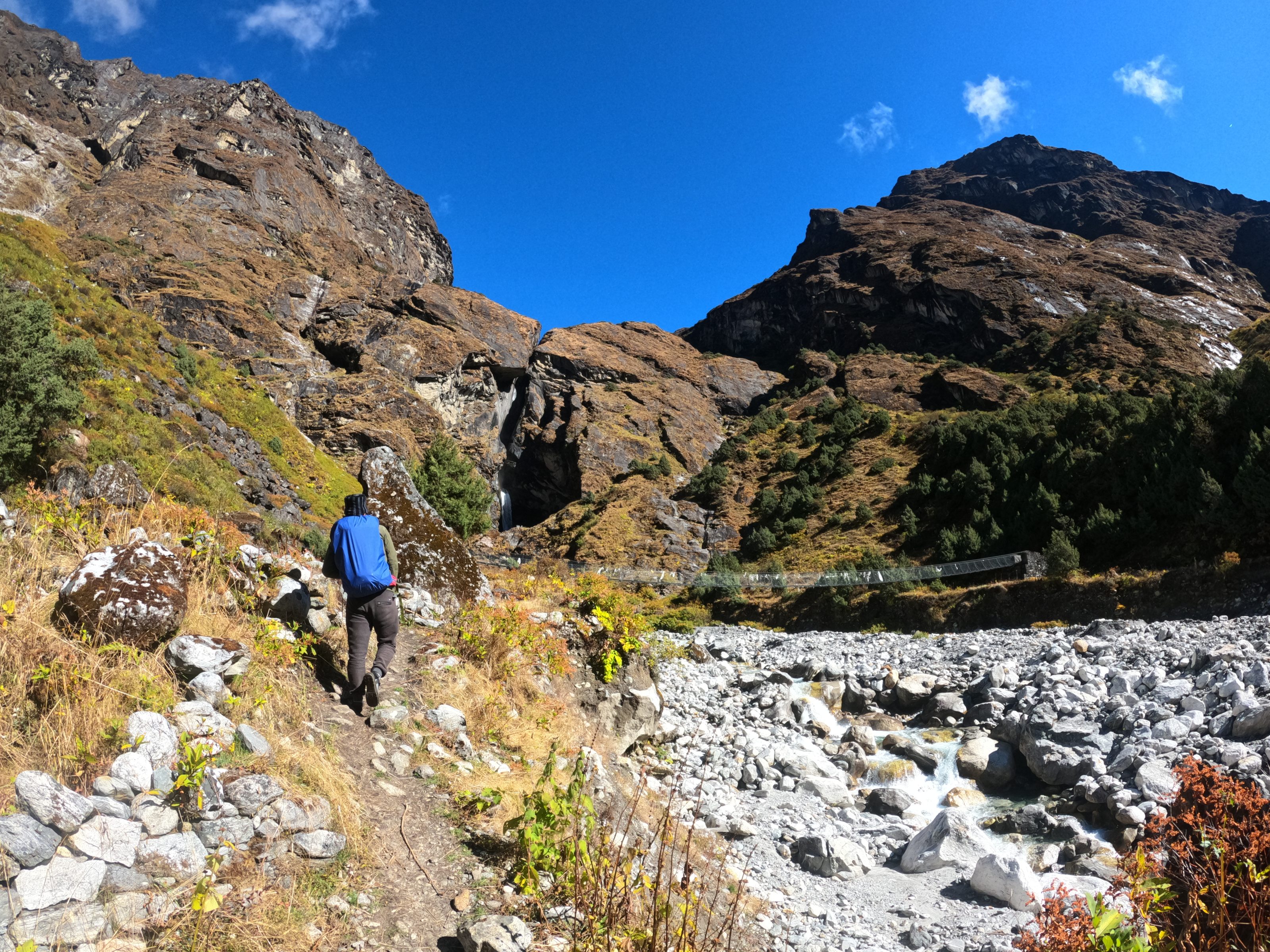 A person wearing a blue jacket and carrying a backpack is hiking on a rocky trail in a mountainous landscape. The scene features rugged mountains with sparse vegetation under a clear blue sky. A rocky riverbed flows alongside the trail, adding to the natural, adventurous setting.