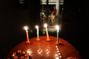 A chocolate birthday cake topped with four lit candles and chocolate decorations, with a softly blurred background showing people in a room.