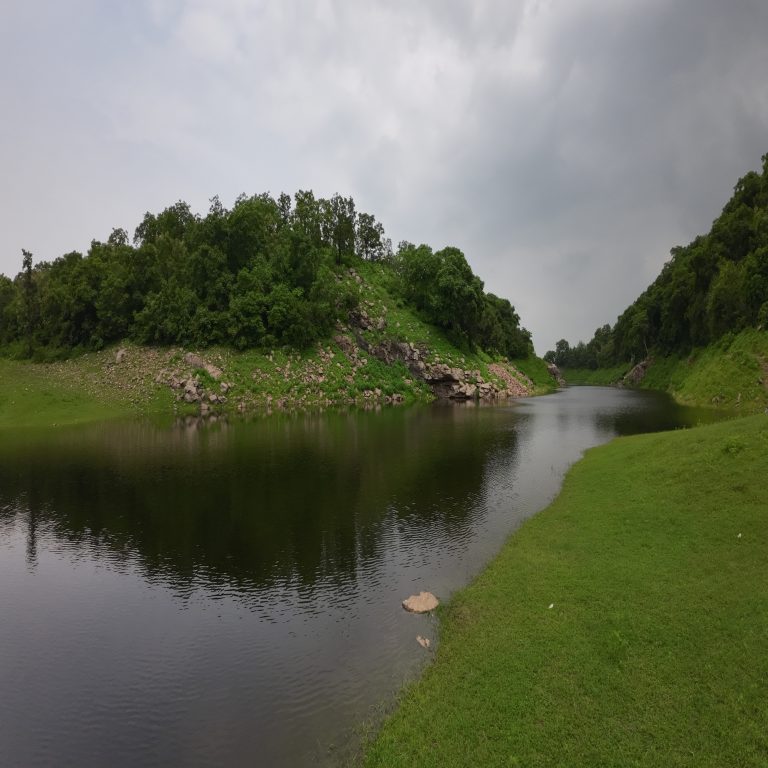 Small lake surrounded by vibrant tree covered green hills, The water reflects the greenery and the cloudy sky above, creating a mirror-like effect. The rocky slope on one side adds texture to the landscape.