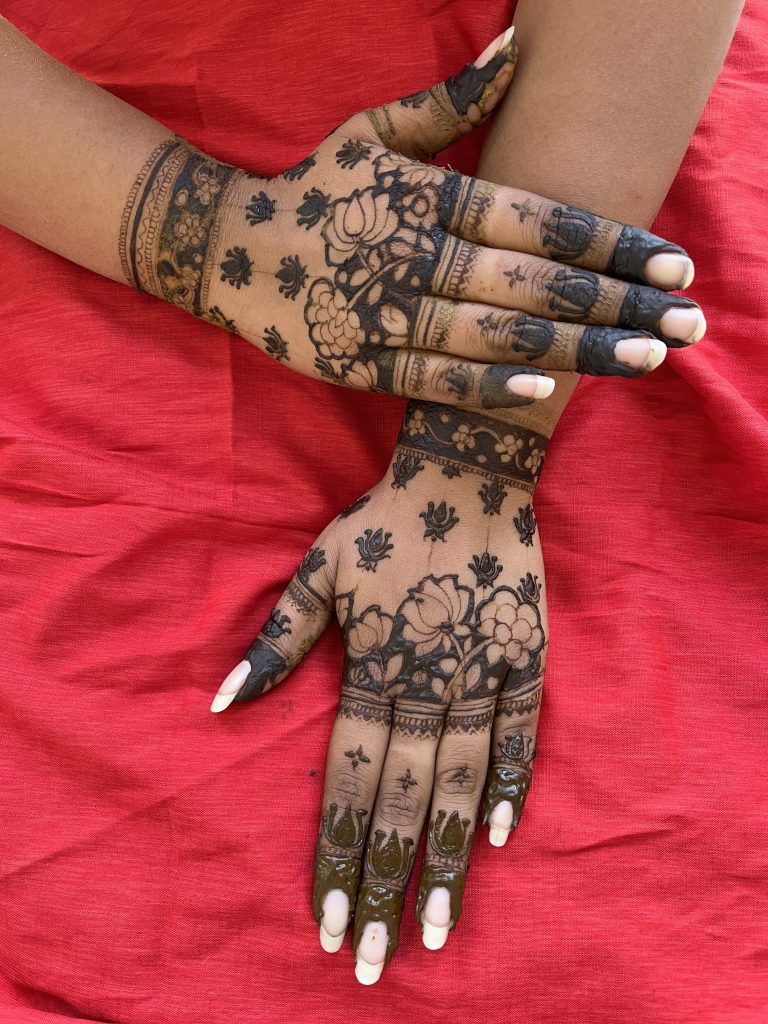 Two hands with intricate henna mendi designs, featuring floral and geometric patterns, placed on a vibrant red fabric background.