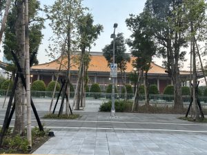 A traditional building with a prominent orange roof is seen behind a row of trees and a modern parking sign. The area in front of the building consists of neatly paved walkways and manicured hedges.