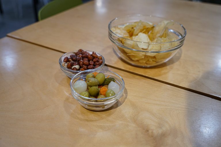 Three glass bowls with snacks on a wooden table: one bowl contains potato chips, another has peeled peanuts, and the third has assorted pickles including olives, onions, and carrots.