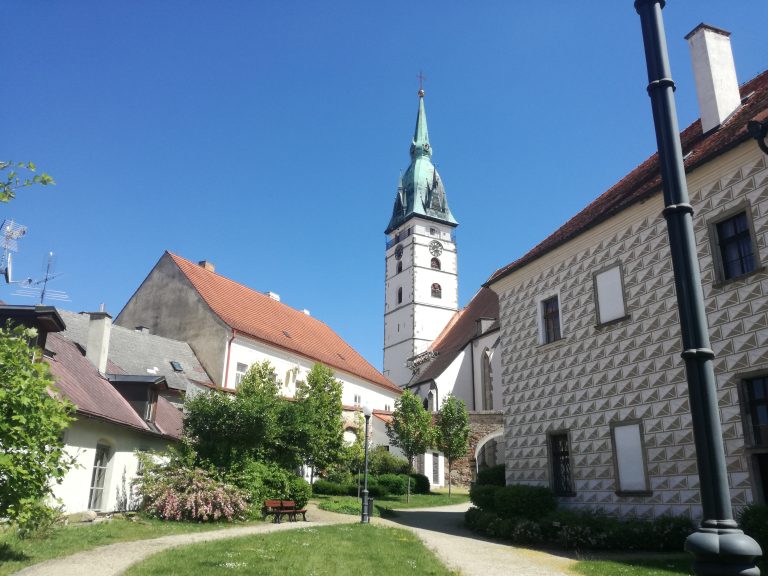 A view of the Church of the Assumption of the Virgin Mary in Jindrichuv Hradec, Czechia. The church features a tall spire with a green roof and clock, surrounded by historical buildings with red-tiled roofs and decorative facades. A small park with paths, trees, and a bench is in the foreground under a clear blue sky.