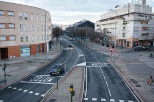 A wide street view featuring two roadways with marked lanes, surrounded by multi-story buildings. A gray car is driving on the left side of the street, while the right lane is reserved for motorcycles, as indicated by the "MOTOS" marking.