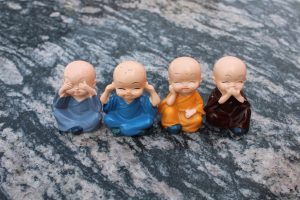 Four small monk figurines in different colored robes are seated in a row on a stone surface. Each figurine is making a gesture representing the "see no evil, hear no evil, speak no evil" proverb, with the fourth adding a playful variation.