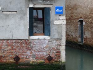 A brick and stone building wall next to a canal. The wall includes a window with open shutters and a blue sign with an icon of a gondola. The waterway is visible, bordered by another brick wall.