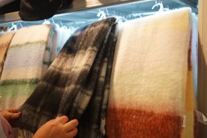 A person is browsing through a selection of soft, multi-colored woolen scarves displayed on hangers in a store. The scarves have various patterns and colors, including stripes and gradient designs. 