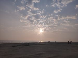 A tranquil beach scene at sunrise, with a partially cloudy sky. The sun is low on the horizon, casting a soft glow over the calm sea. Two small boats are silhouetted in the distance, and a few people are walking along the beach