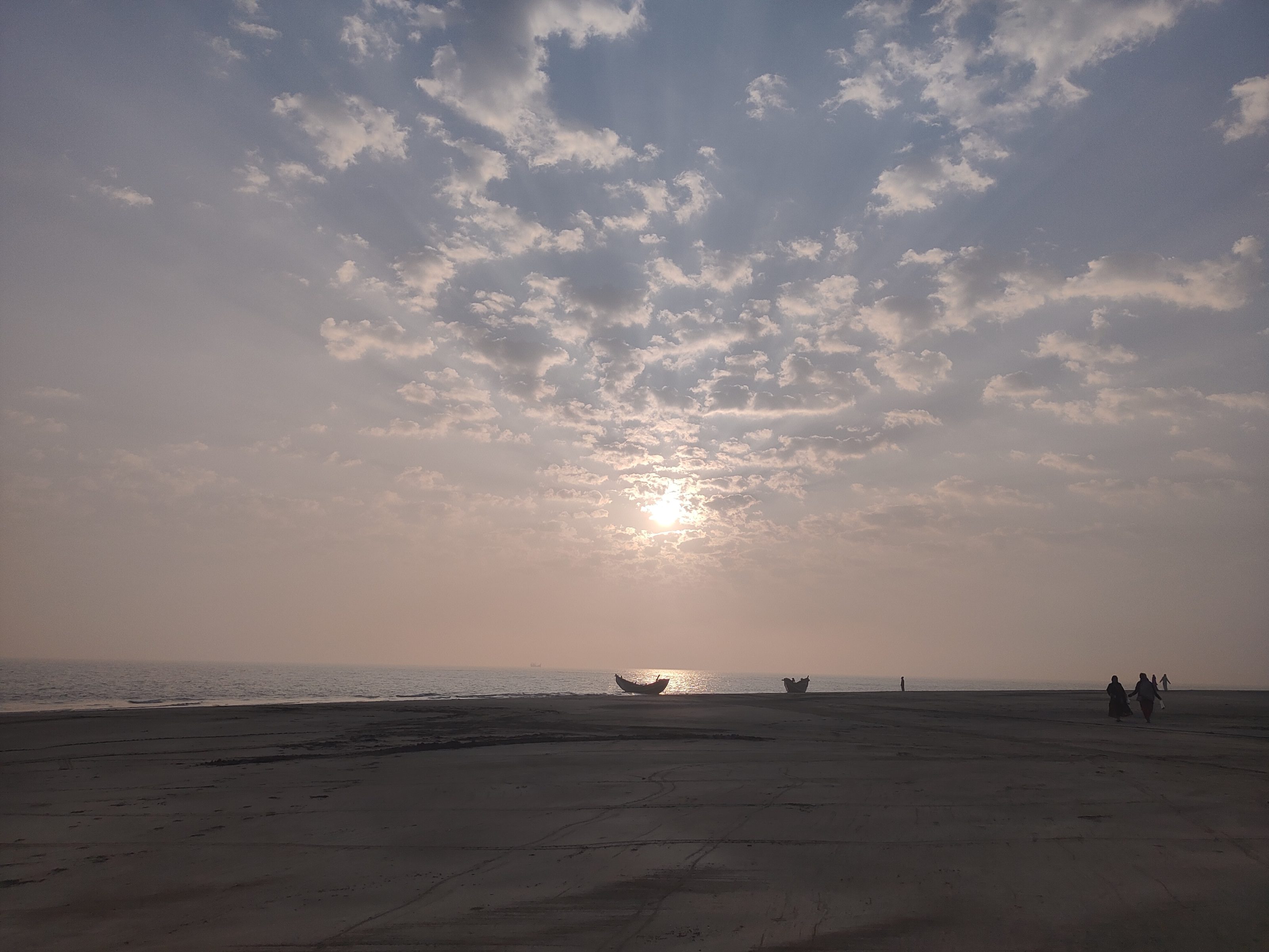 A tranquil beach scene at sunrise, with a partially cloudy sky. The sun is low on the horizon, casting a soft glow over the calm sea. Two small boats are silhouetted in the distance, and a few people are walking along the beach