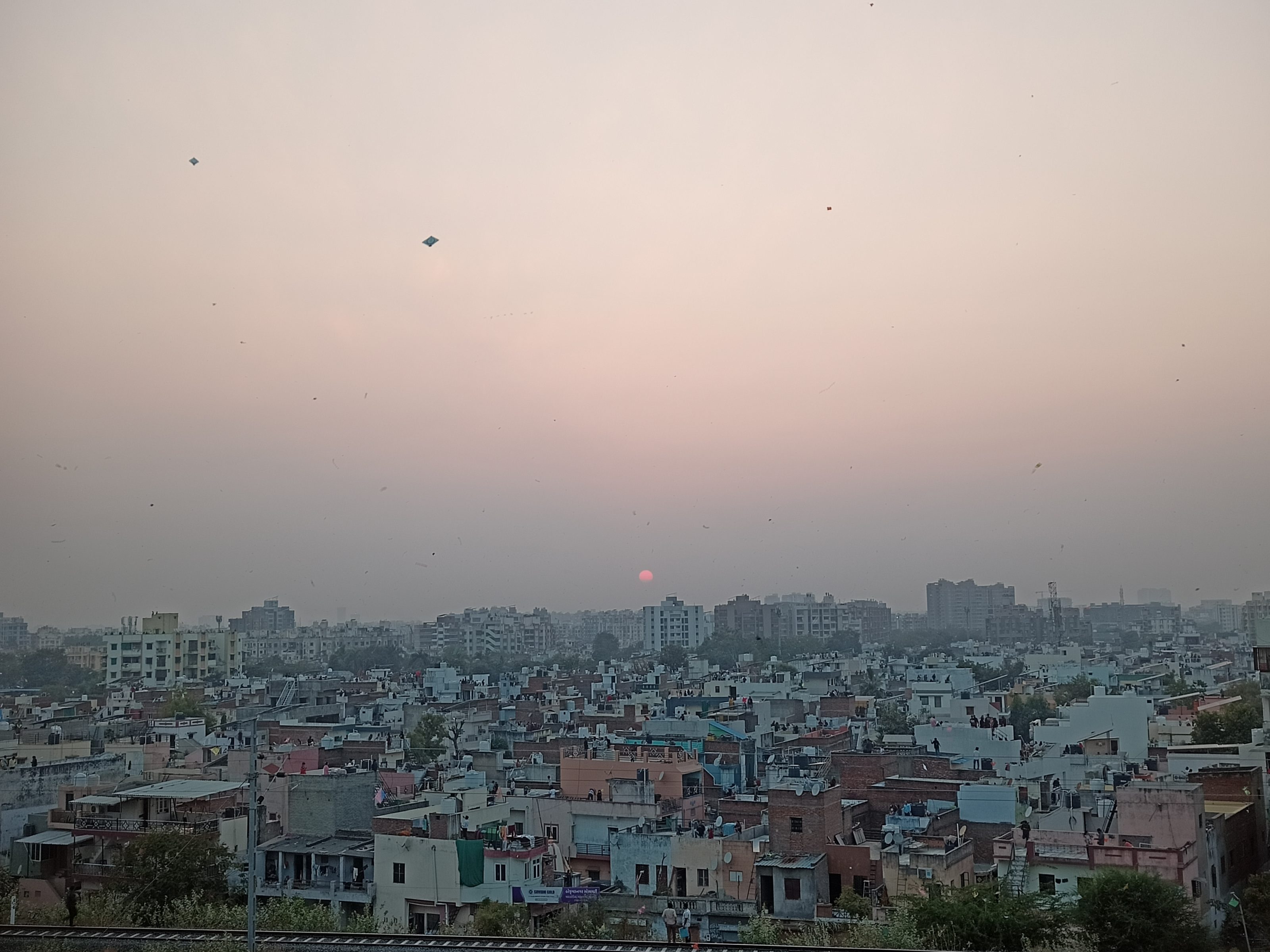 Captivating cityscape at sunset with a light blue sky transitioning to orange hues. Kites gracefully soar in the sky, adding a festive charm, while the glowing orange sun creates a warm and serene ambiance.