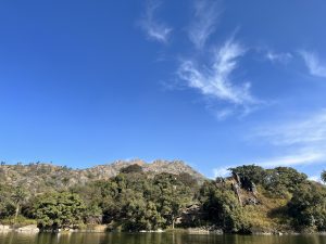 A scenic view of a clear blue sky over a lush green forested hillside with rocky outcrops. A small body of water is visible in the foreground, reflecting the greenery and sky above