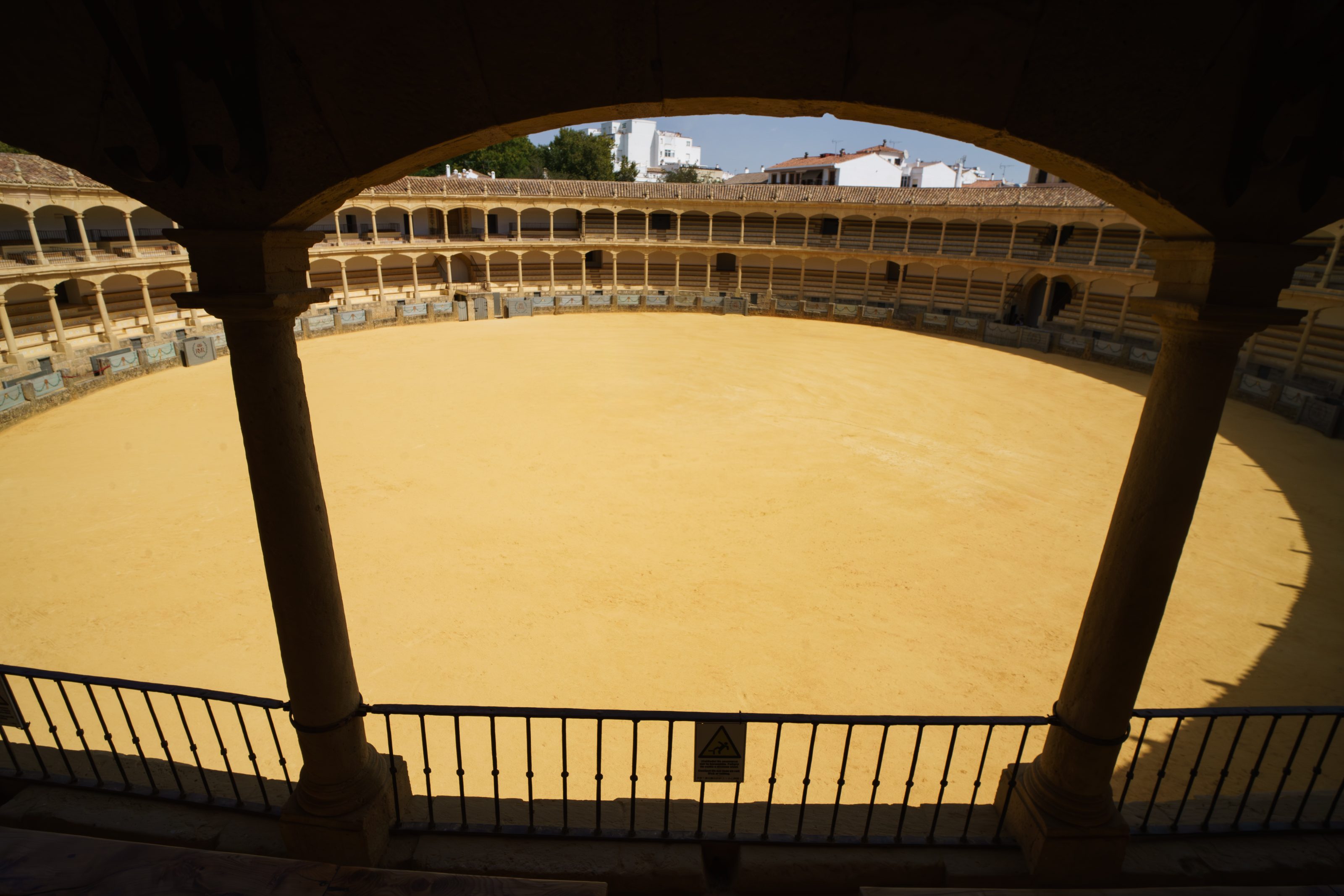A view of a large empty bullring arena, circular sandy ground surrounded by tiered seating and arched columns. The architecture is traditional, with warm beige tones. Terracotta roofed white building beyond the arena