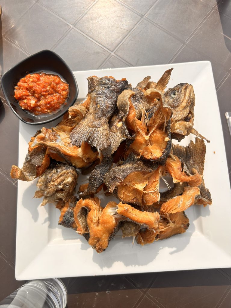 A plate of fried fish pieces served on a white square plate, accompanied by a small bowl of red chili sauce on the side.