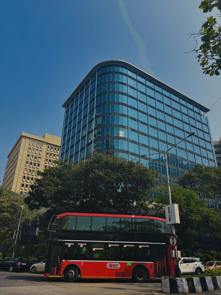 A red and black double-decker bus is driving on a city street with modern glass buildings and leafy trees in the background under a clear blue sky.